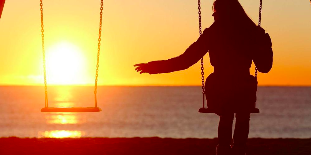 Silhouette d'une personne assise sur une balançoire en bord de mer au coucher du soleil, tendant la main vers une deuxième balançoire vide, avec une lumière dorée représentant le deuil.